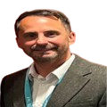 A picture of Stephen Bartholomew. He is wearing a grey suit jacket over a white shirt with a blue lanyard around his neck. He is looking toward the camera and smiling.
