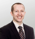 An image of Steve Tebbutt, MHA. He is smiling toward the camera wearing a black suit jacket with a white shirt and a brown, blue and white striped tie.